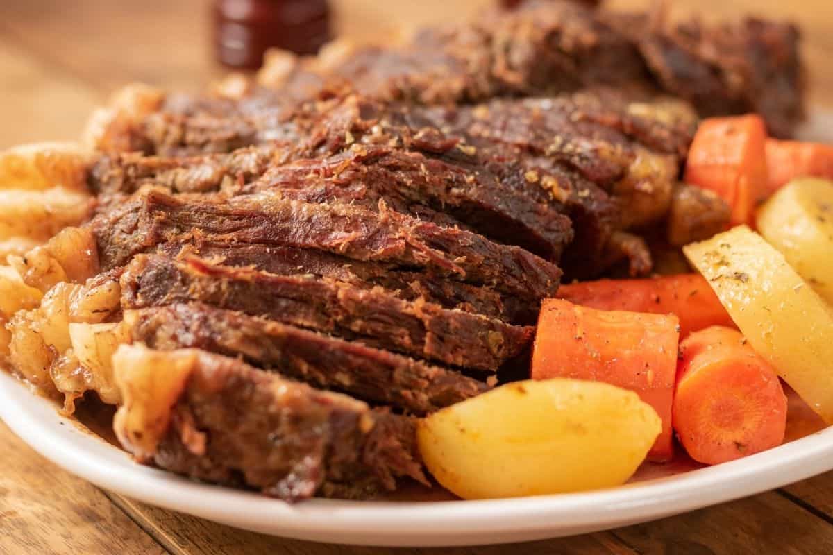 Close up of a sliced beef pot roast surrounded by onions, potatoes and carrots on a white plate on a wooden tabletop.