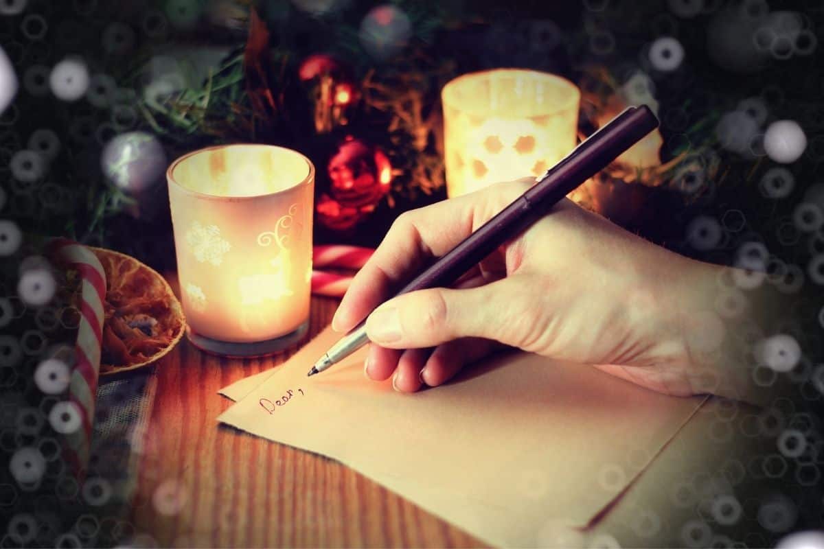 A right hand writing on two pieces of brown paper on a wooden desk with dried orange slices, candy canes, candles, and fresh pine boughs