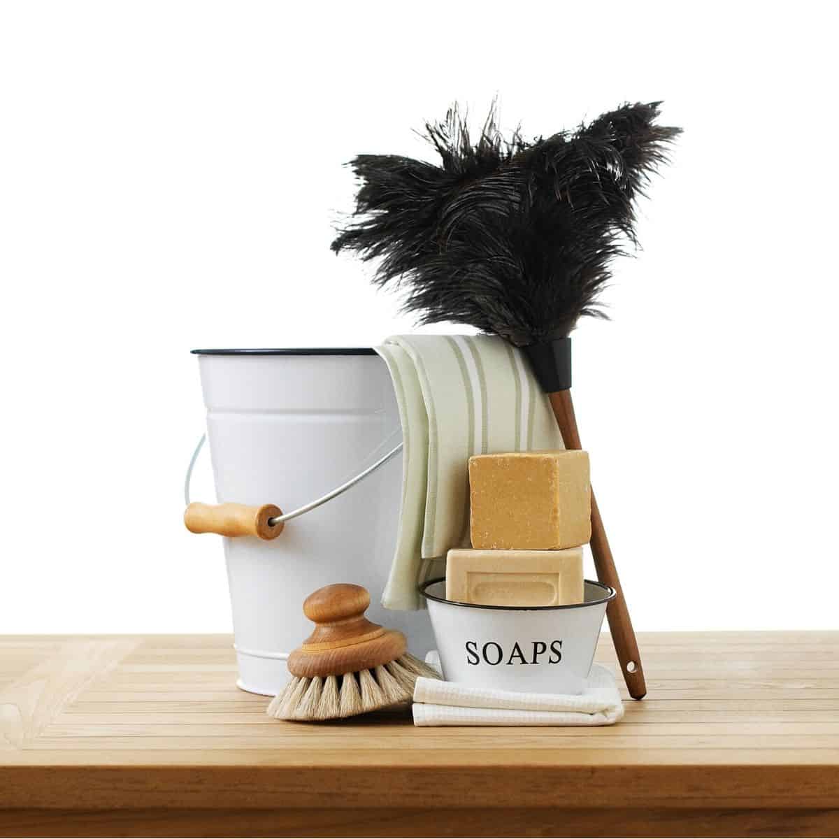 White bucket with wooden handle, striped towel draped over the side of the bucket with black feather duster, wooden scrub brush, and tin with homemade soaps stacked inside.