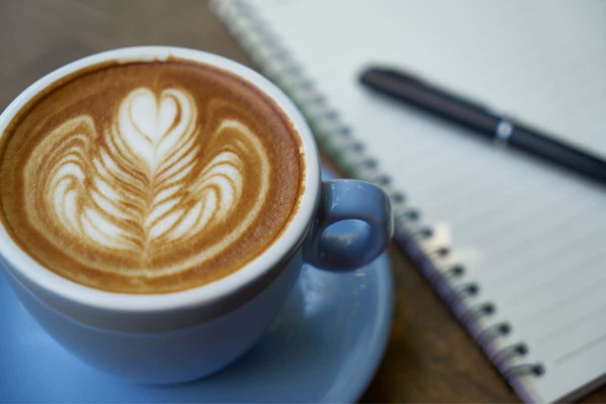 Mug with a latte with flower design in foreground. Pen and paper fuzzy in background.