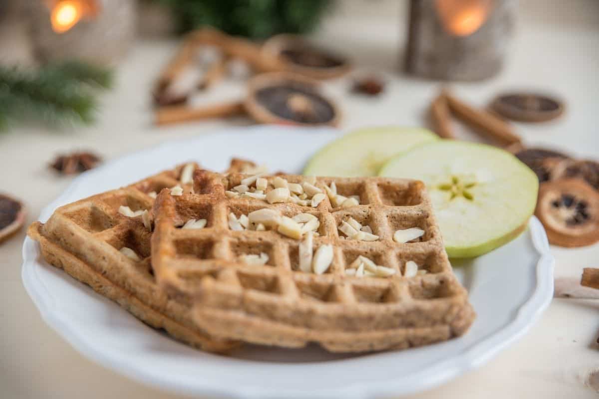 Two waffles topped with almonds on a plate with apple slices. Plate is surrounded by dried orange slices, pine sprigs, and lit candles.