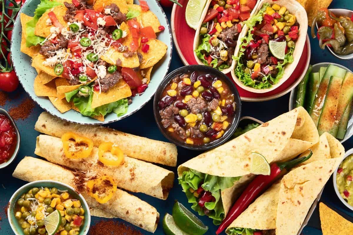 A Mexican Feast with nachos, taco salad, black bean soup, tortillas, soft shell tacos, taquitos, and small dishes with peppers, corn, and lime.
