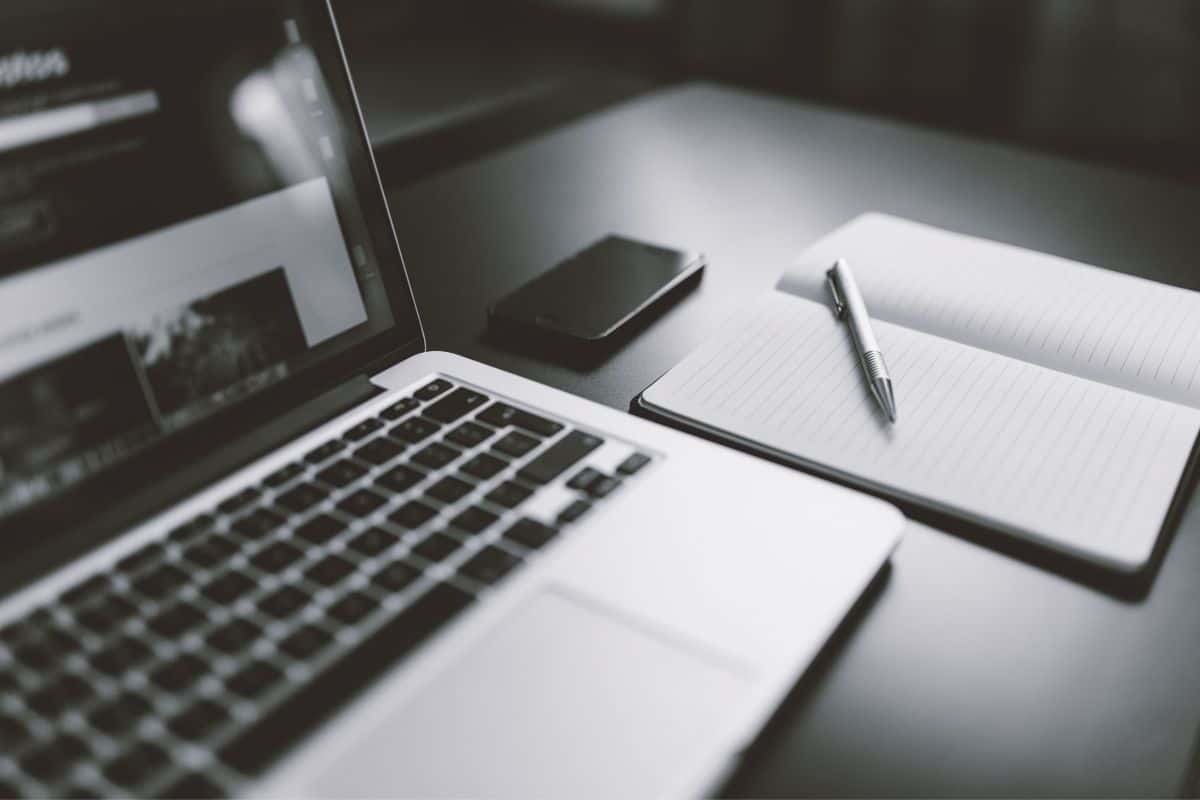 Black and white photo of an open laptop, a notebook and pen, and a smartphone on a shiny black tabletop.