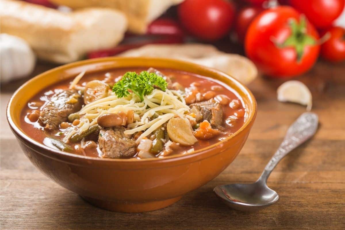 Brown bowl of soup with spoon resting next to it on wooden tabletop. Whole tomatoes and loaves of bread in the background.