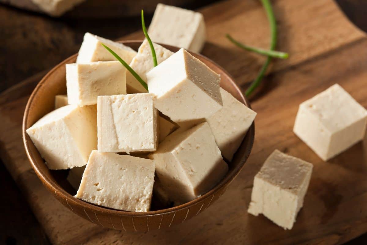 brown bowl of chopped raw tofu sitting on a wooden cutting board, garnished with green onions