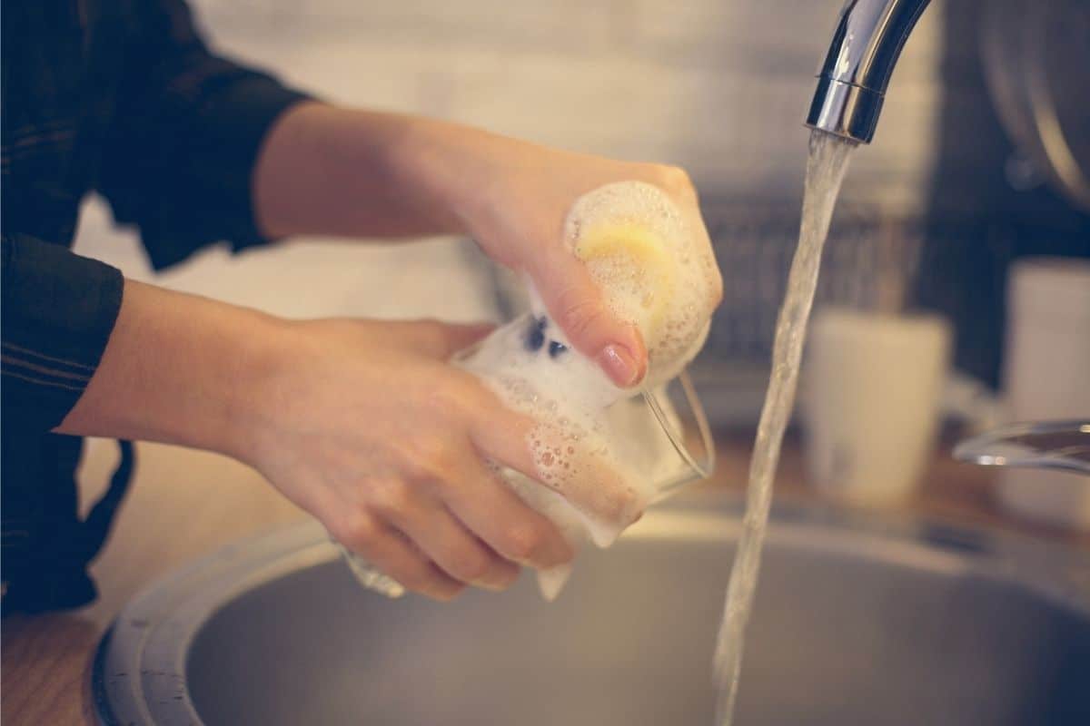 A woman in a black shirt is washing dishes in a small round sink. Her hands only are visible along with the streaming water from the faucet and the soapy glass she's washing with a yellow sponge.