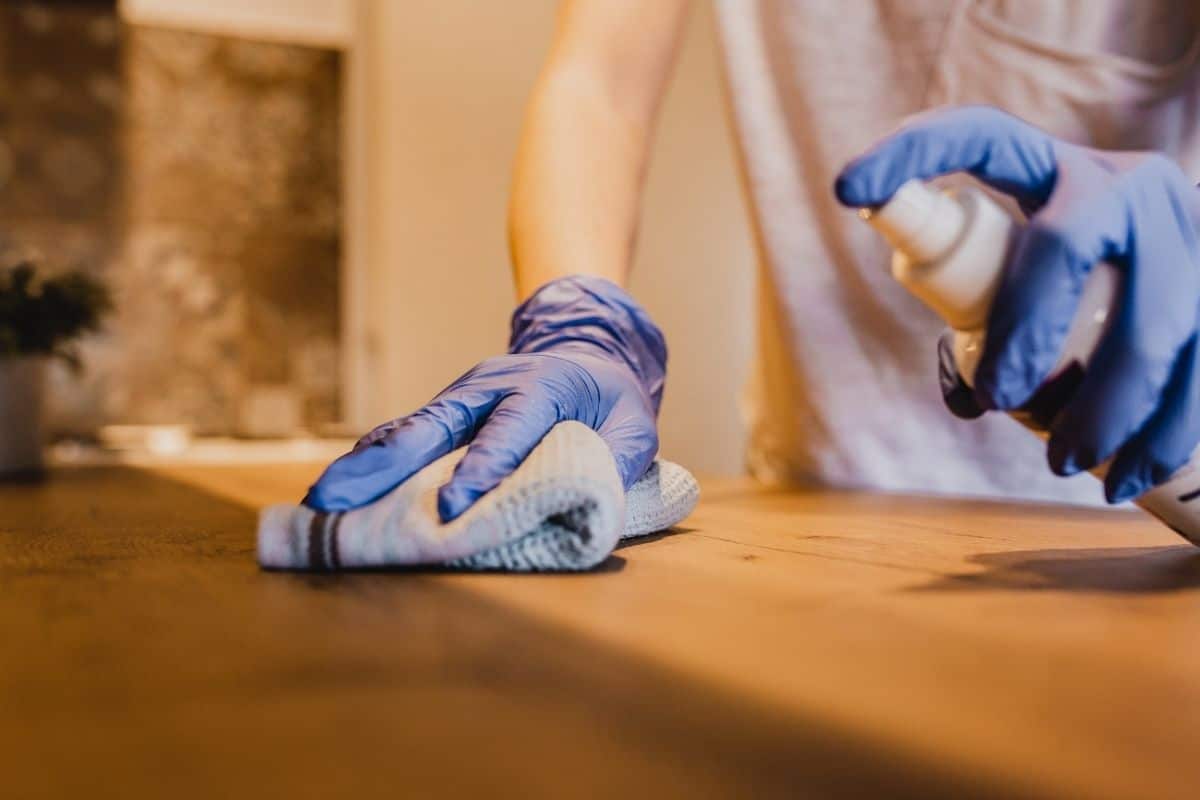 A woman in a gray shirt is polishing a wooden tabletop. She is using a spray and a cotton cloth and is wearing blue latex gloves.