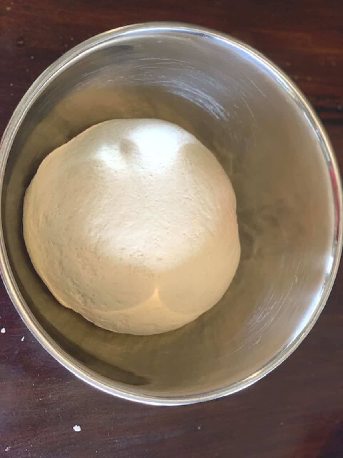 Bread dough in a stainless steel bowl on a dark wooden tabletop