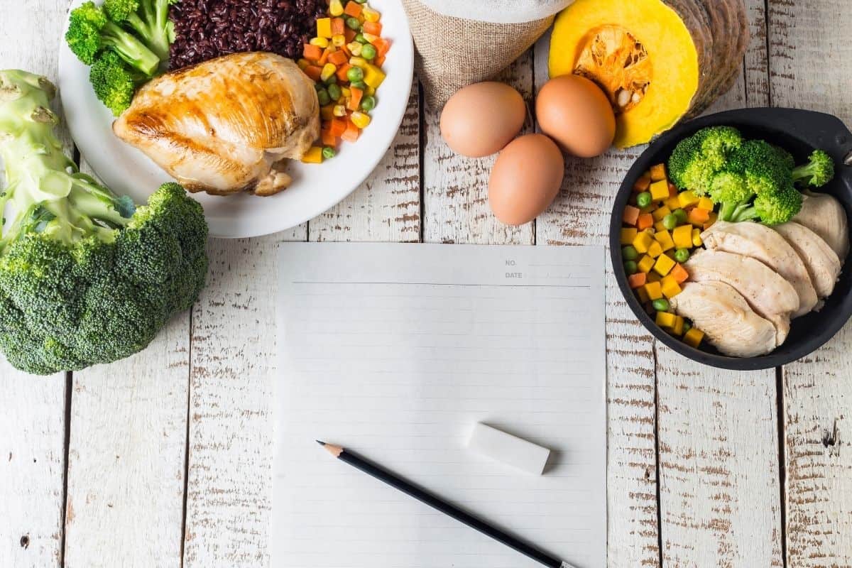 Broccoli, a dish of chicken, eggs, squash, and a pan of veggies and meat on a wooden tabletop surrounding a piece of paper with a pencil and eraser on it.