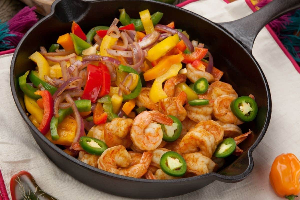 A cast iron pan resting on a dish towel filled with shrimp and yellow bell peppers, jalepenos, and red onions. 
