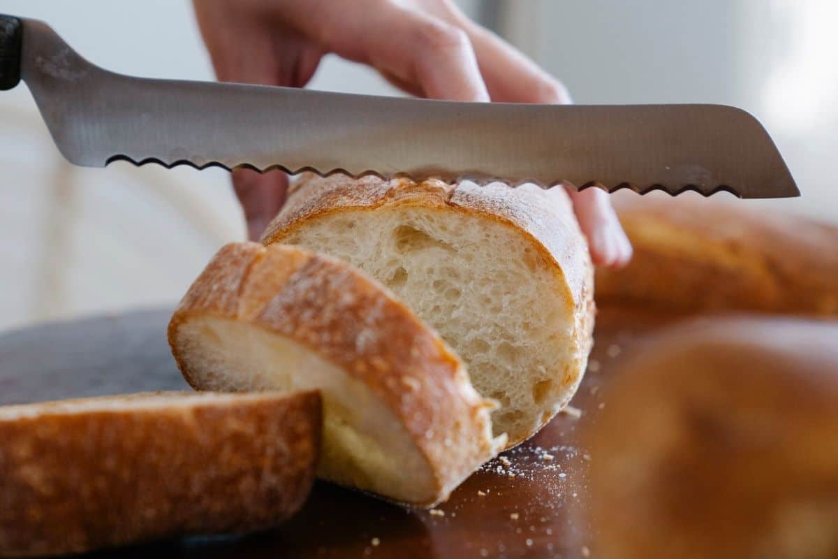 A pair of hands cutting a baguette with a serrated bread knife.