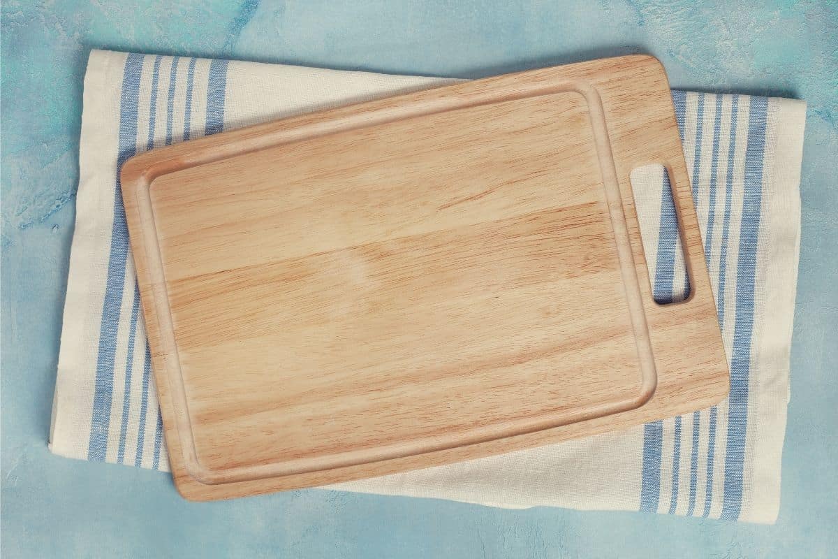 A rectangular wooden cutting board with a handle resting on a blue and white linen tea towel on a blue countertop.