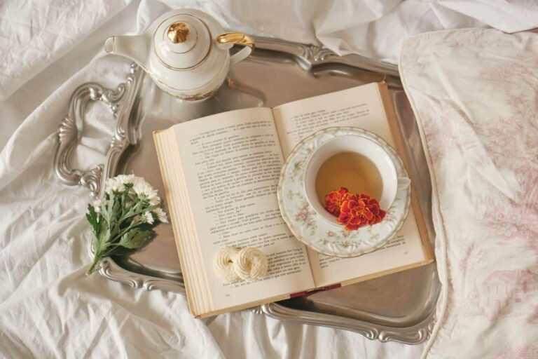 Silver tray on a white bedsheet with a teapot, cup of tea, flowers, and an open book.