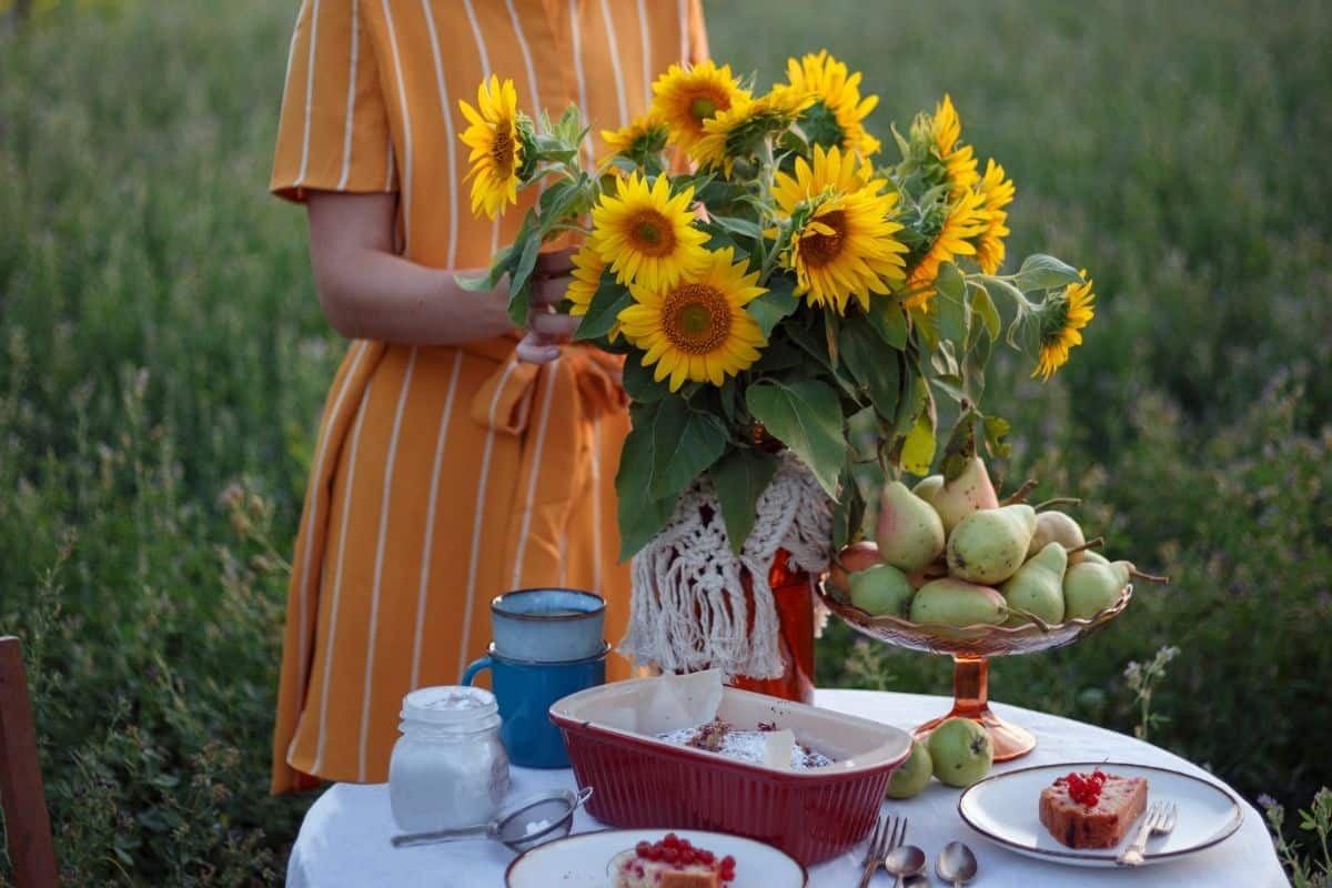 A girl in an orange dress in a field of grass setting a small tea table with a vase of sunflowers. The table is filled with sweet things to eat, silverware, and mugs.