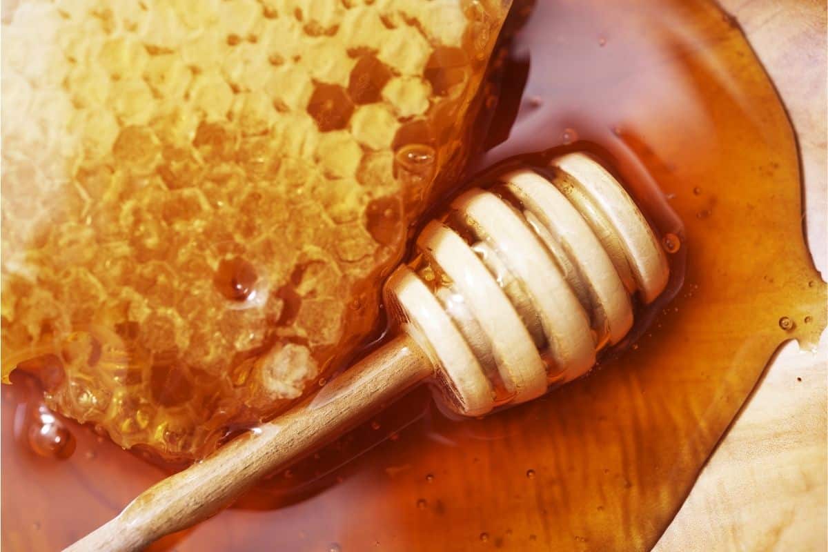 A wooden honey dipper and a honeycomb resting in a pool of honey on a wooden surface.