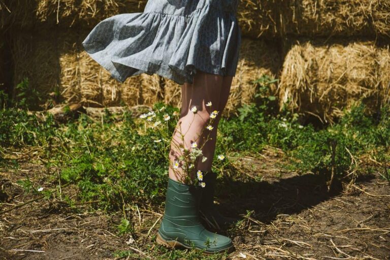 A girl in a blue skirt and rubber boots standing in a muddy barnyard in front of haystacks