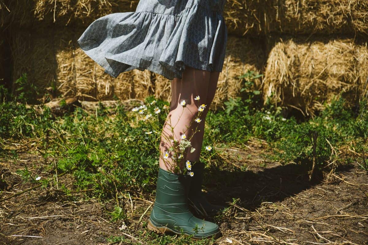 A girl in a blue skirt and rubber boots standing in a muddy barnyard with haystacks in the background.
