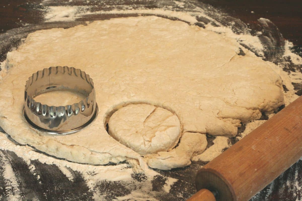 Biscuit Dough rolled out on a floured surface. Biscuit cutter resting on the top of the dough, rolling pin in the front.