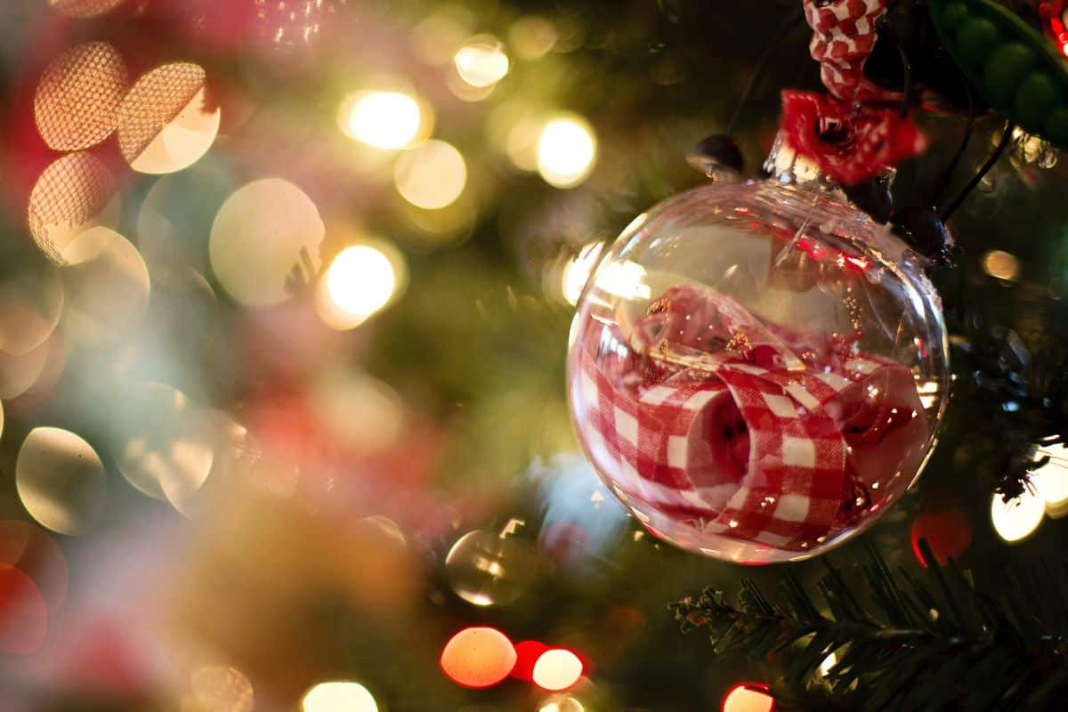 A glass ornament filled with red and white gingham ribbons hanging from a Christmas tree with white blurry Christmas lights in the background