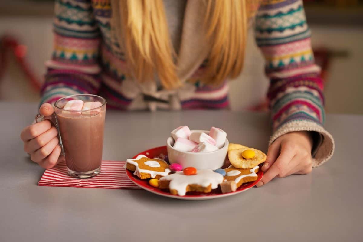 A girl with long strawberry blonde hair in a decorative Christmas sweater sitting at a table in front of a cup of hot cocoa and a plate full of Christmas cookies and sweets