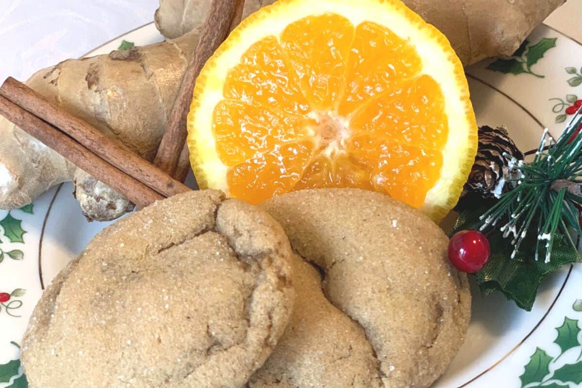 Two soft ginger sourdough cookies on a Christmas patterned plate with a ginger root, cinnamon sticks, an orange slice, and a small holly branch.