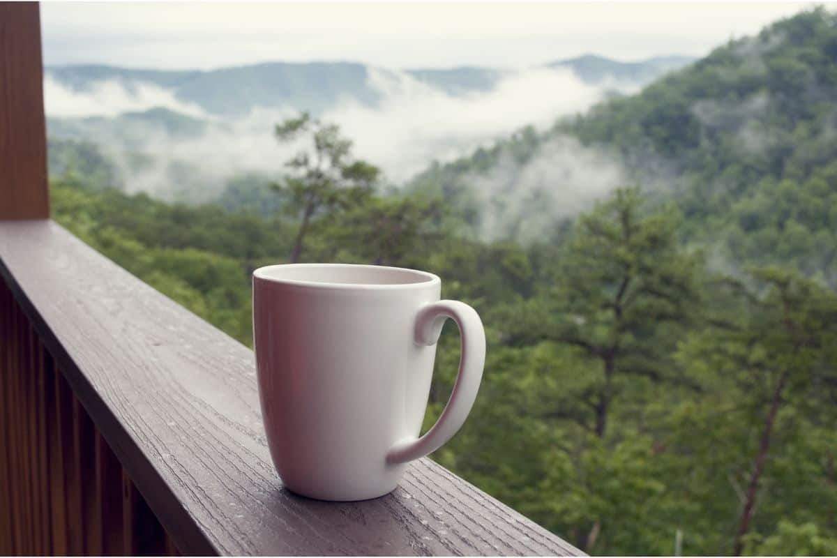 A white mug sitting on a wooden rail overlooking a mountain scene with clouds resting on the mountaintops.