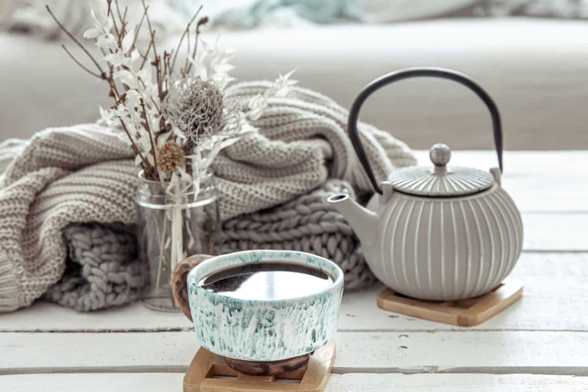 A gray teapot with a black handle, a chunky knit sweater, a mason jar filled with twigs and flowers, and a full teacup on a wooden coaster all sitting on a weathered white coffee table.