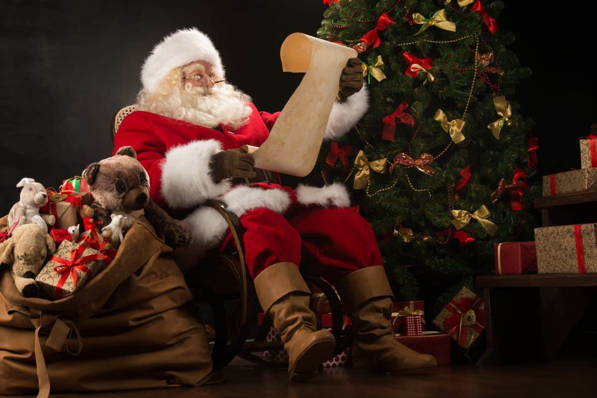 Santa Claus sitting in a caned rocking chair next to his sack of toys and a decorated Christmas tree.