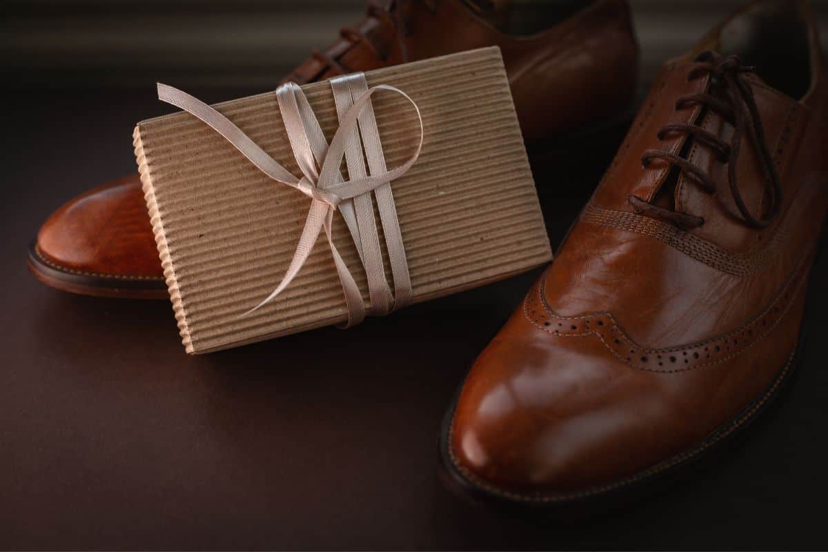 A pair of men's brown dress shoes sitting on a dark brown surface with a brown gift box wrapped in cream ribbon sitting between the shoes.