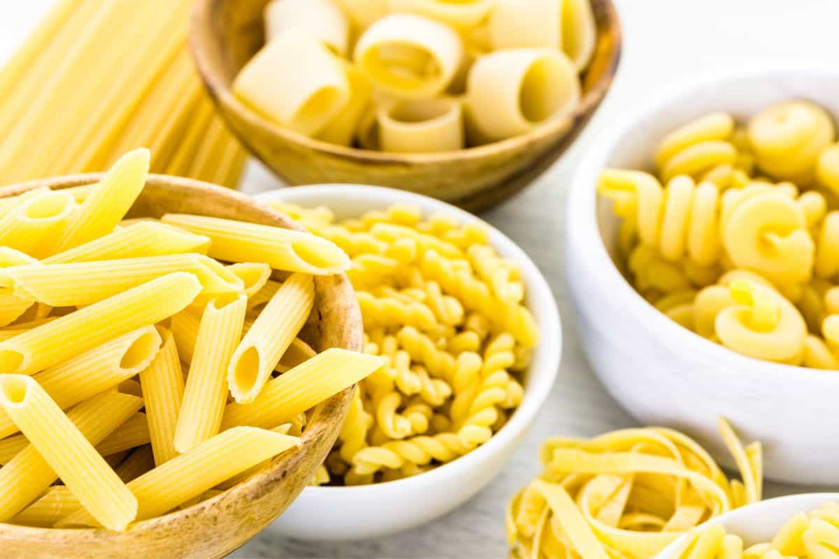 Different types of dry pasta in white and wooden bowls and lying directly on a white marble countertop.