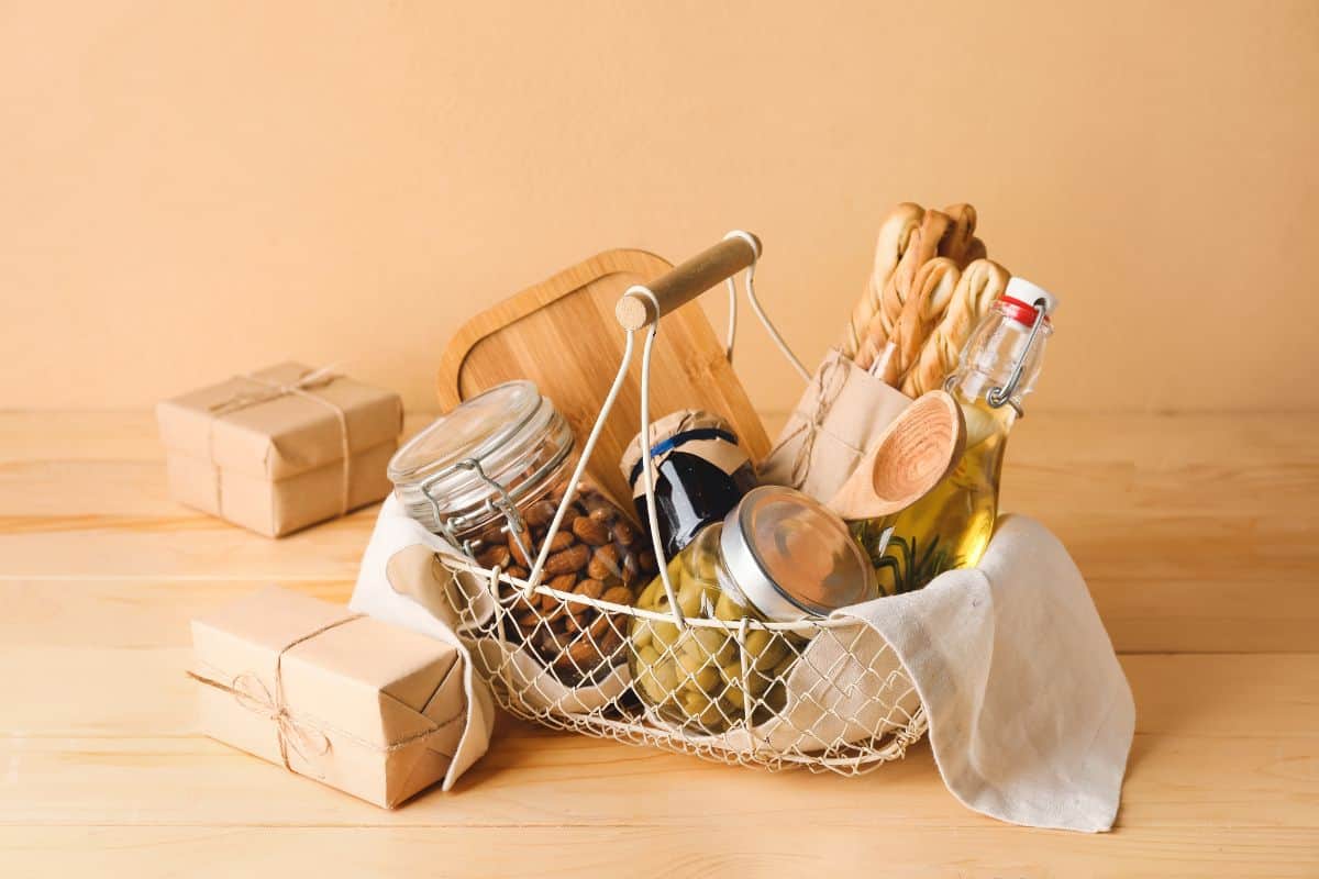 White Wire Basket with wooden handle filled with a wooden spoon and cutting board, bread sticks, a glass bottle of oil, and jars of olives, almonds, and jam. The basket is sitting on a wooden surface next to two gifts wrapped in brown paper and twine.