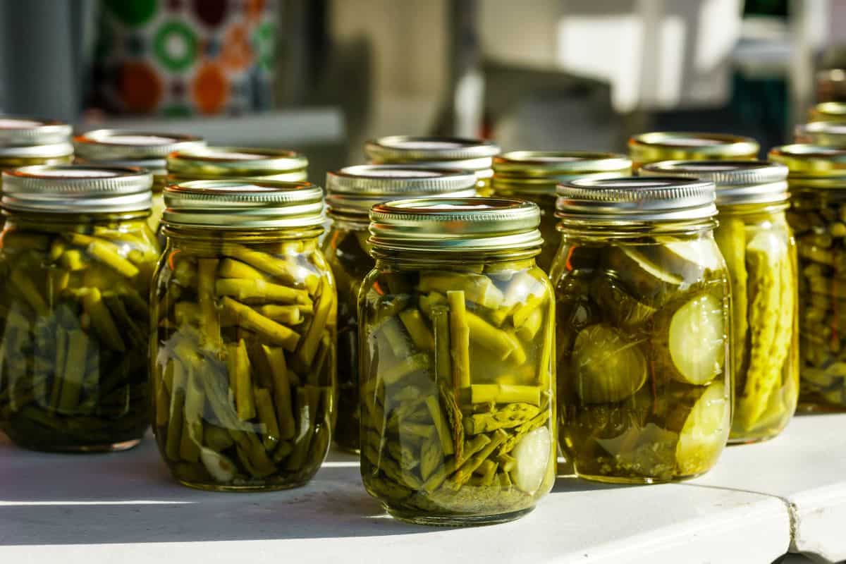 Home canned mason jars full of green beans, pickles, cucumbers, and other green vegetables on a white tabletop
