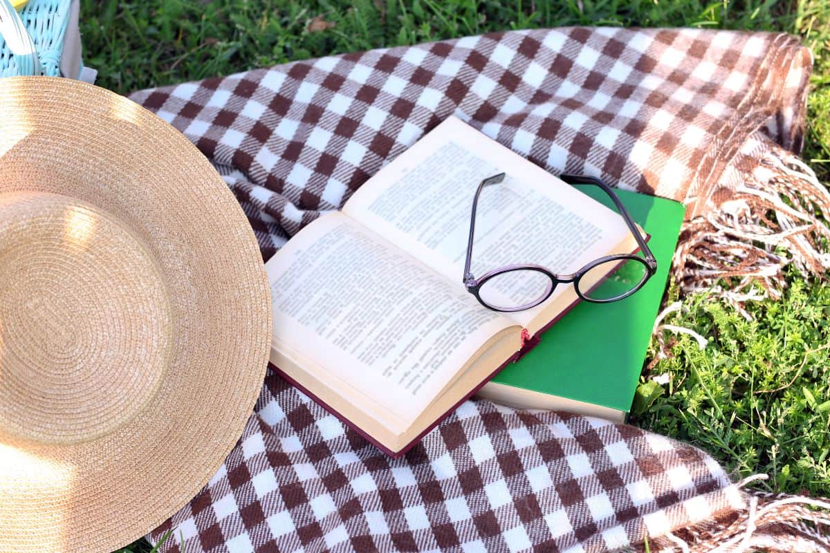A brown gingham blanket lying on the grass with a straw hat and two books, one lying open with a pair of glasses on top.