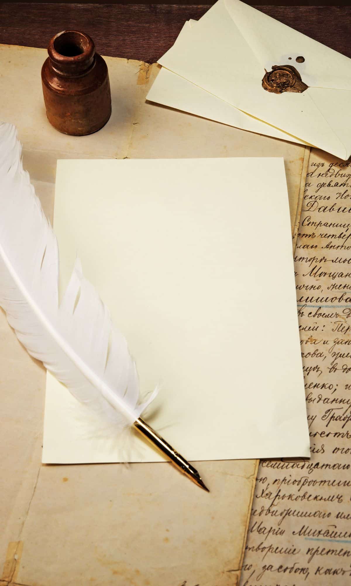 Piece of paper lying open on top of antiqued paper with an ink bottle and feather quill lying on top. Above the parchment are two envelopes, one of which has a wax seal