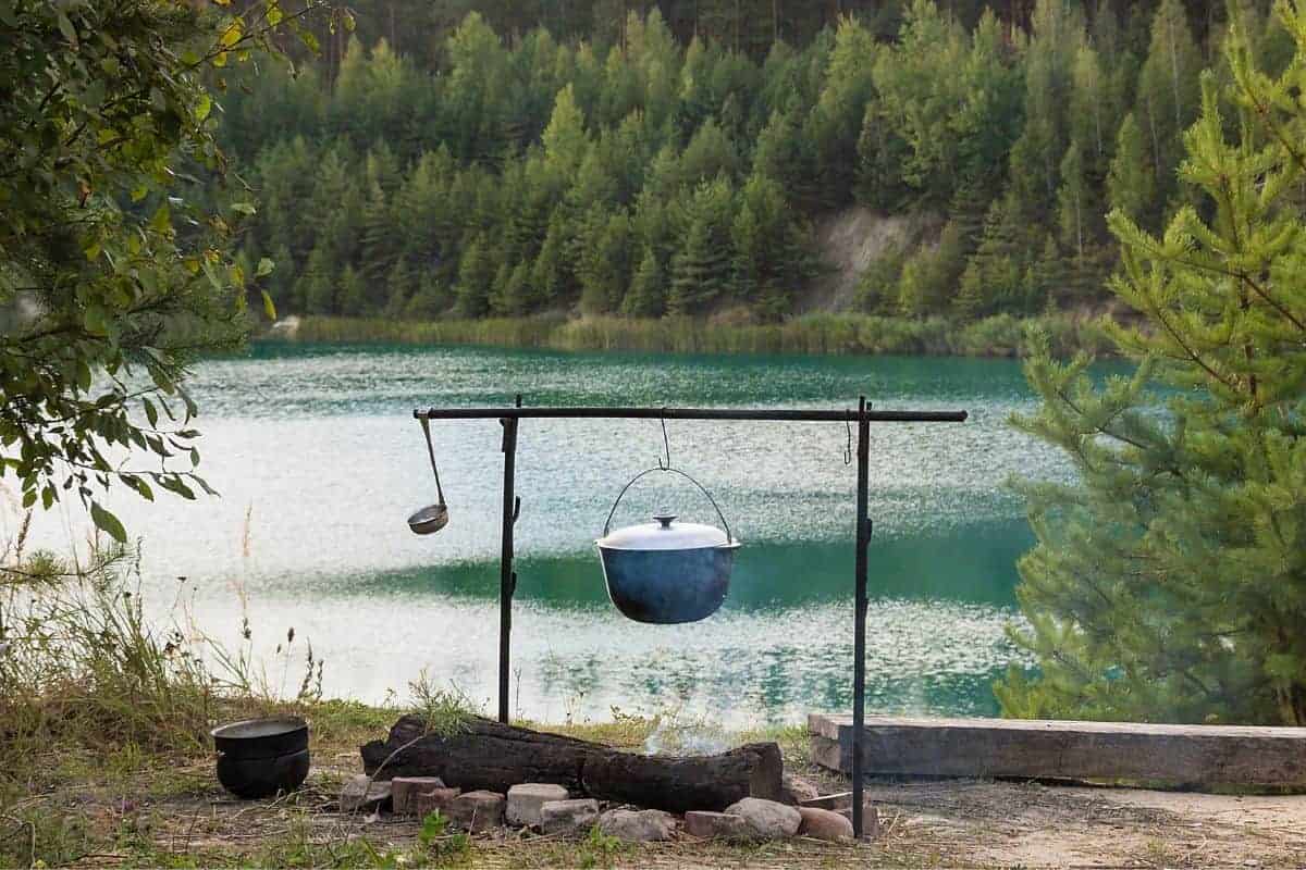 A cast iron pot hanging over a fire with a ladle hanging on the end of the pole. There's a lake and a mountain in the background.