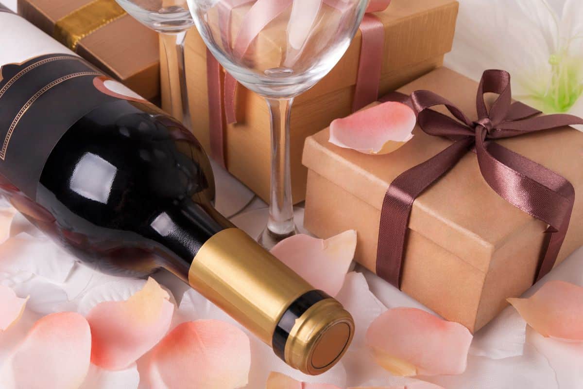 Bottle of wine lying on flower petals next to two wine glasses and three brown paper wrapped gifts