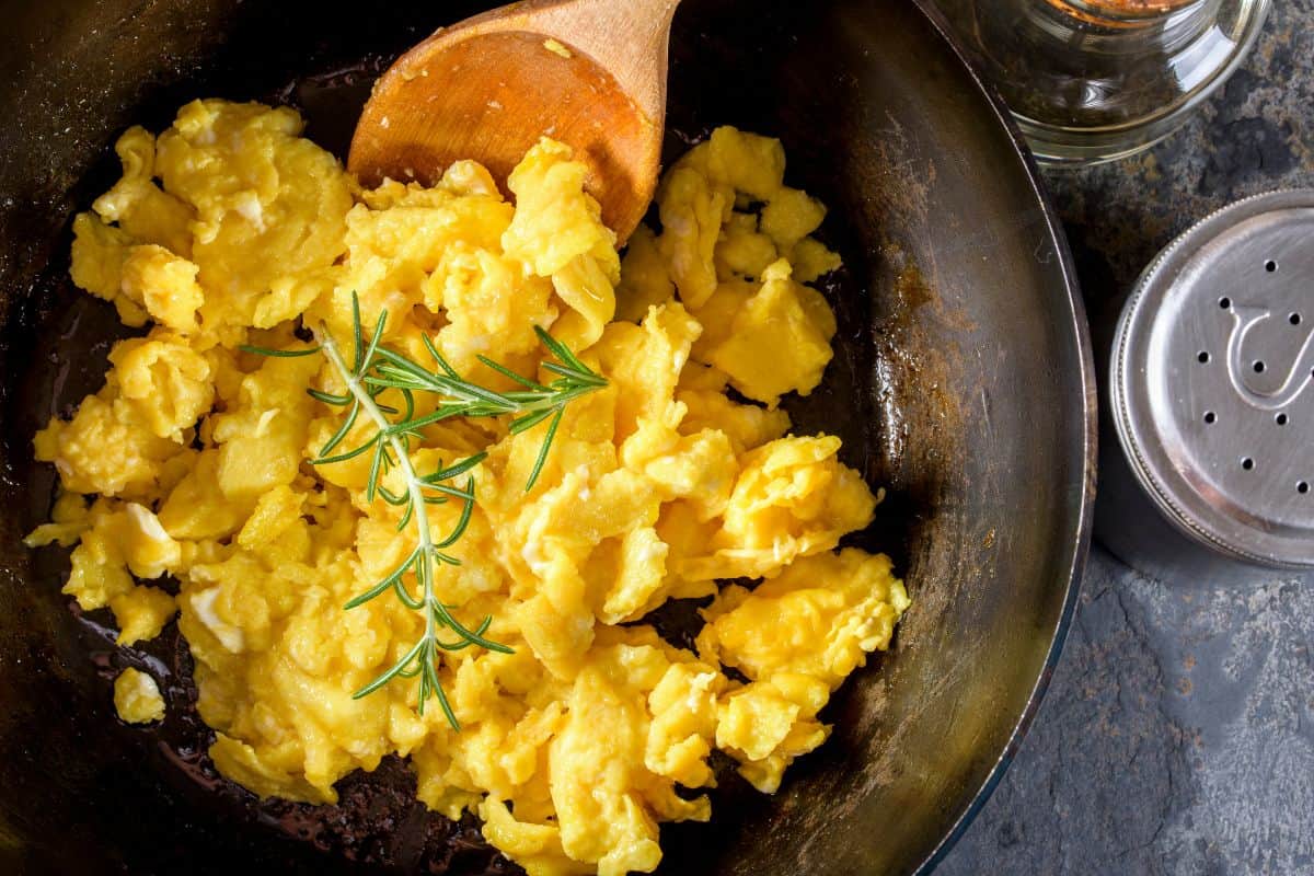 Scrambled Eggs in a cast iron skillet topped with fresh rosemary being stirred with a wooden spoon